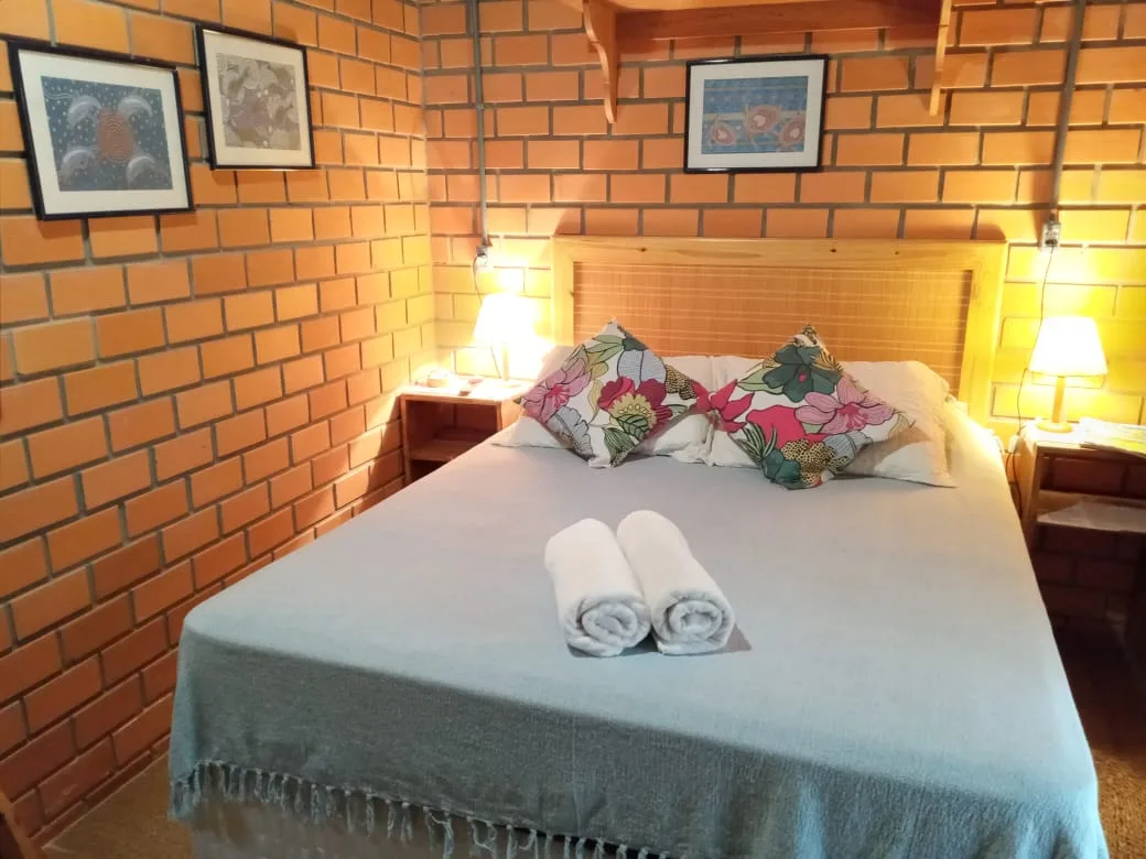 Cama de casal com cabeceira de palha e quadros na parede de tijolinho da suíte