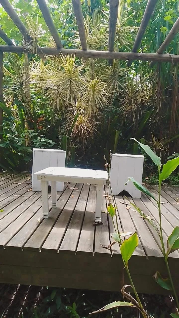 Deck de madeira do Espaço Zen com mesinha e bancos brancos sob o pergolado, cercado de plantas