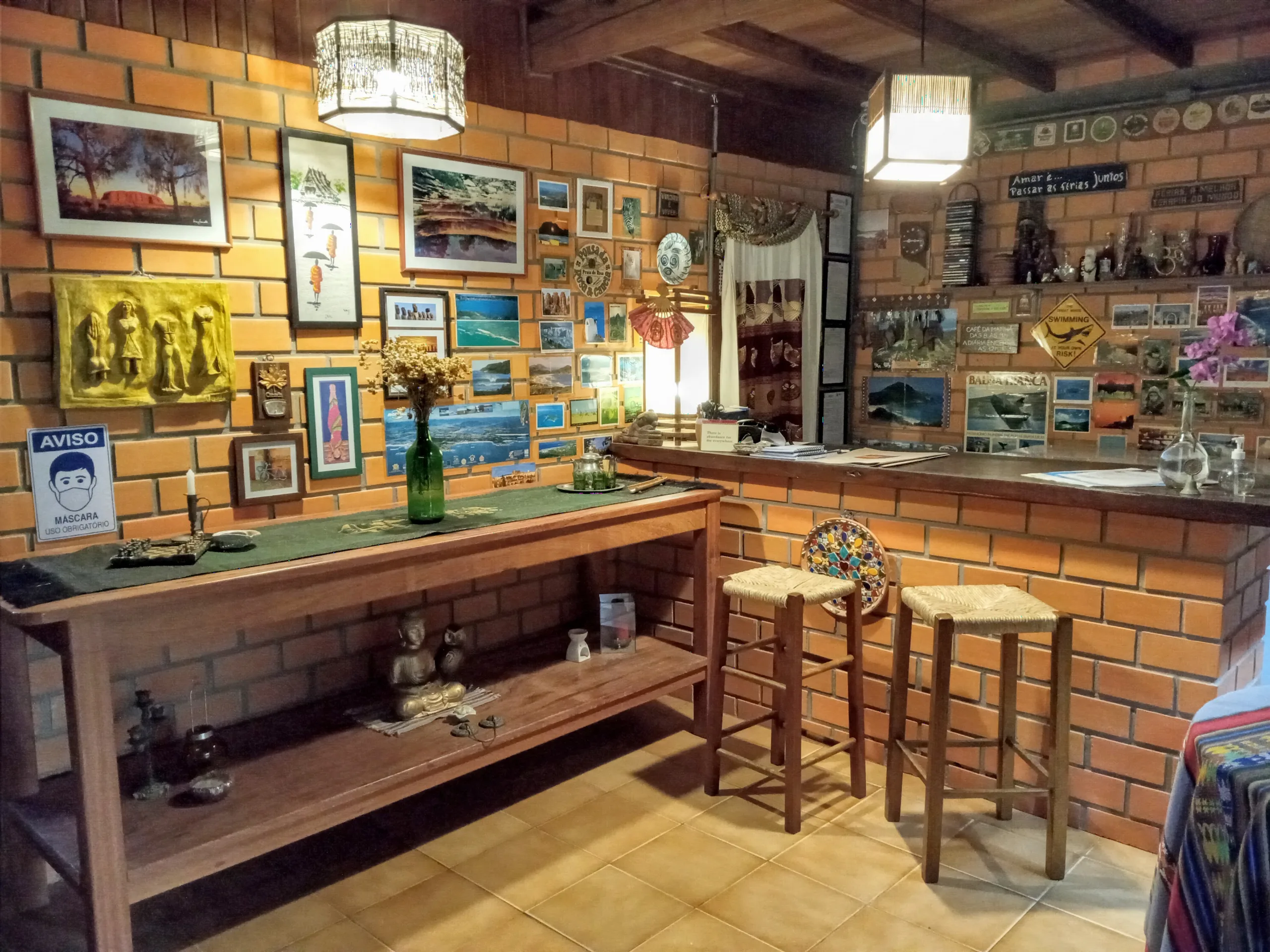 Recepção e bar da pousada com balcão de tijolinho, banquetas, luminárias e quadros da Praia do Rosa